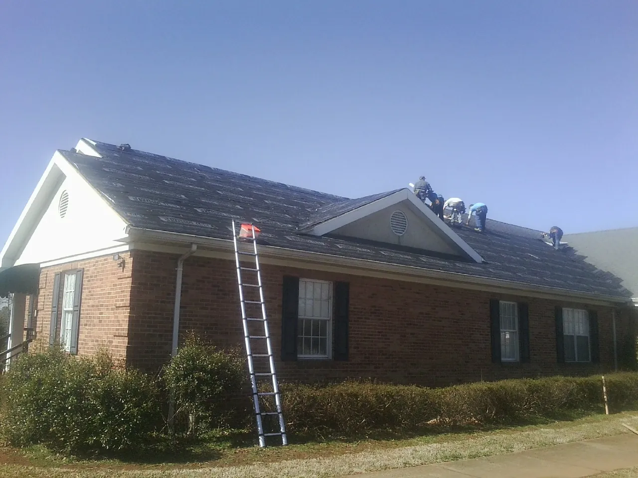 Crew tearing shingles off a brick ranch roof with a ladder set — Metal Roof Repair work by our crew in Corrigan